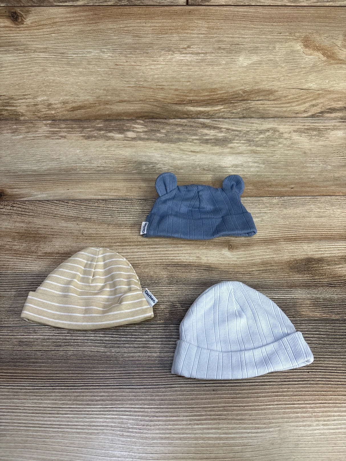 Flat lay of three Gerber newborn hats: blue hat with ears, beige striped hat, and white cuffed hat on wood background