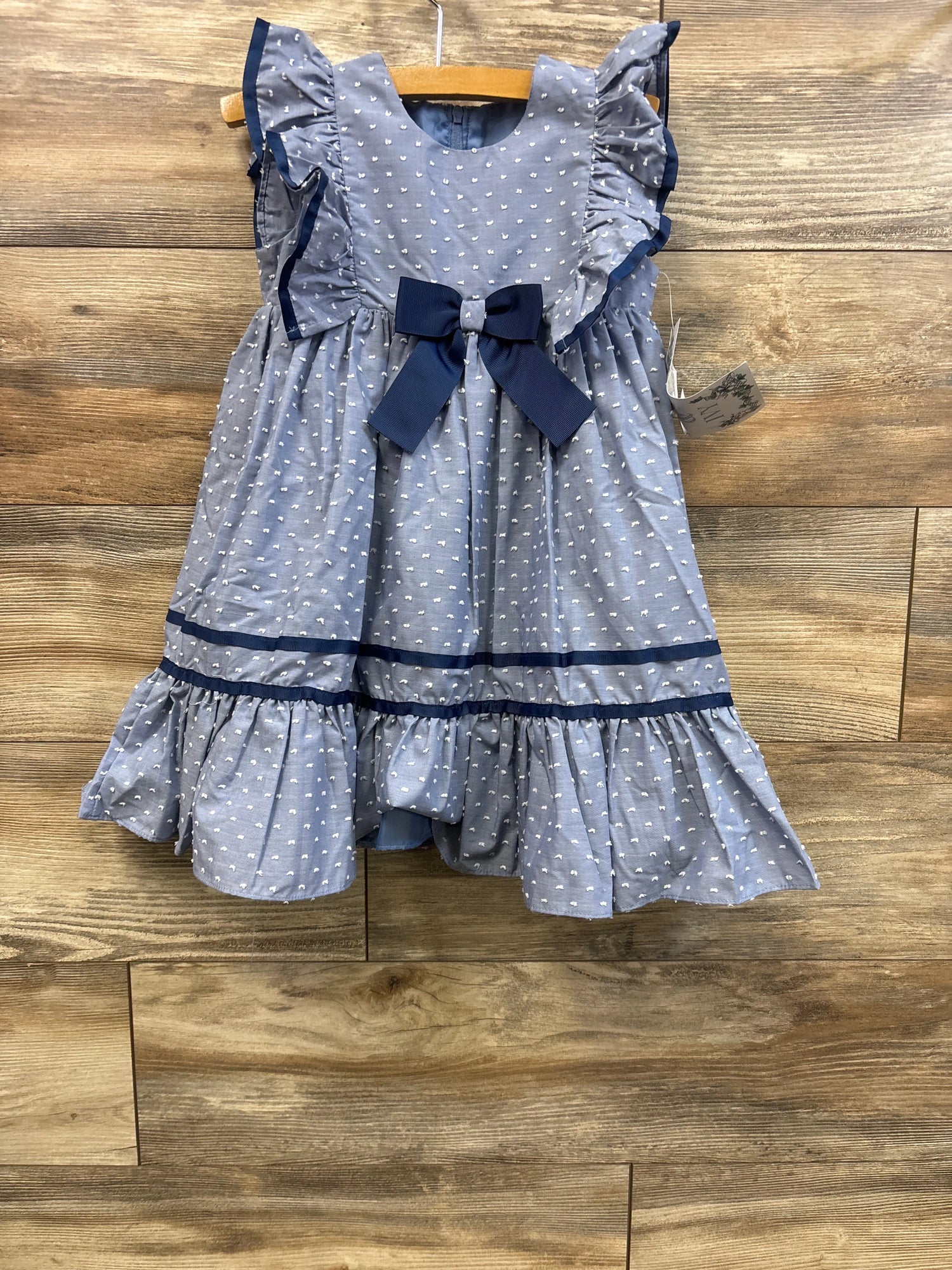 Front view of Iris & Ivy Swiss dot dress in blue showcasing ruffled sleeves, navy bow, and tiered ruffle hem on wood floor