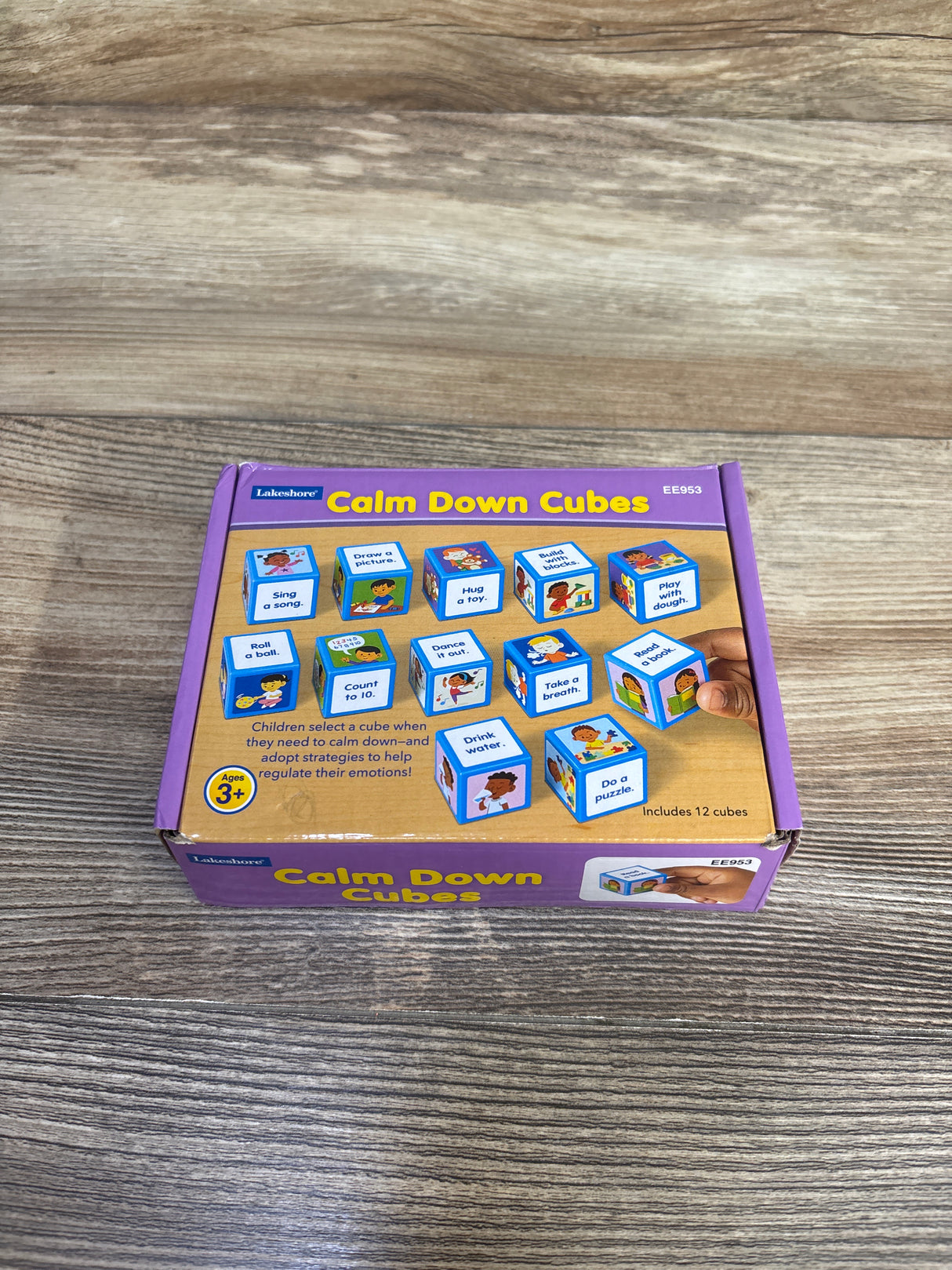 Top view of Lakeshore Calm Down Cubes box showcasing illustrated activity cubes and a hand holding a cube on a wood tabletop