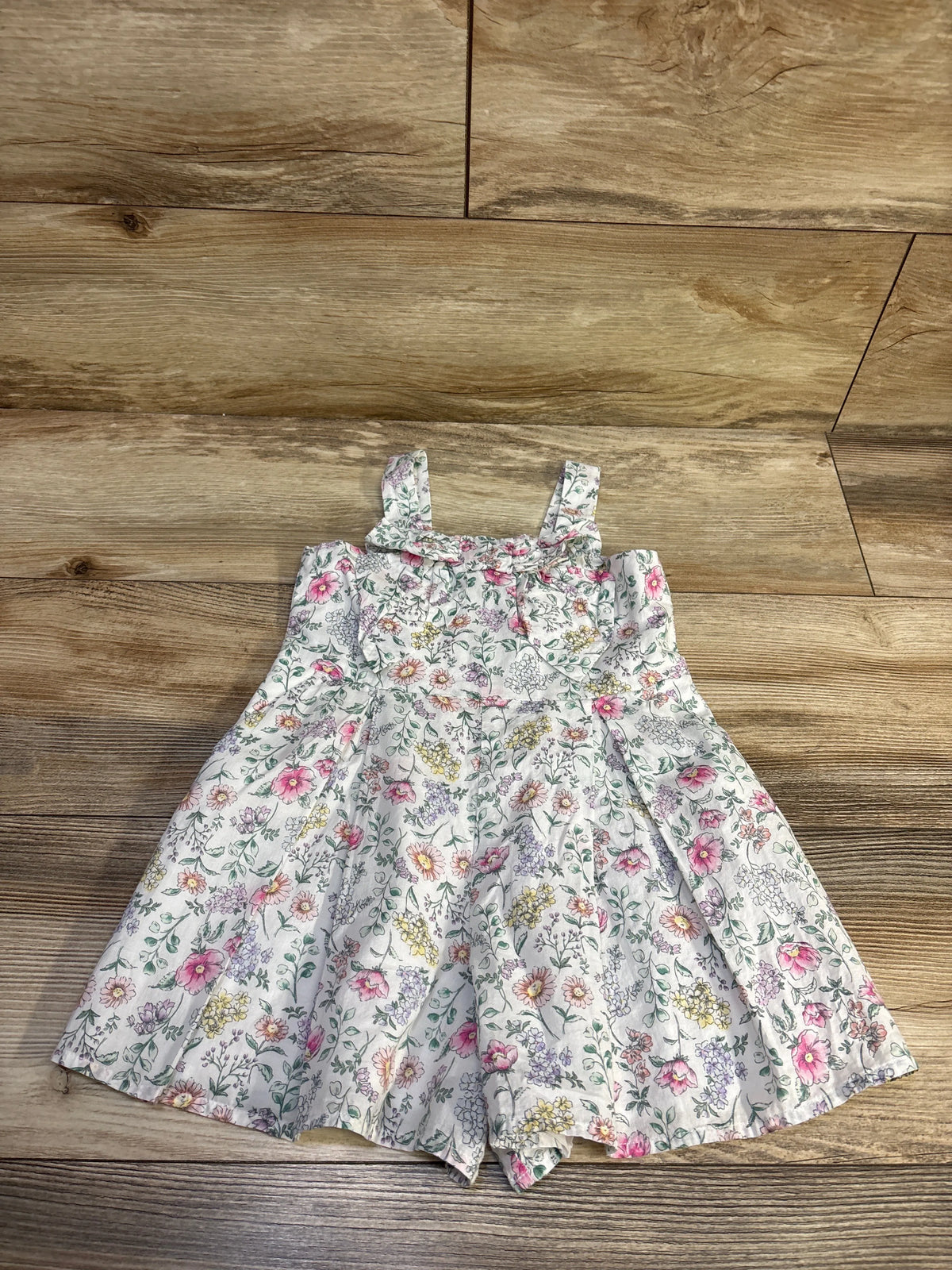 Front view of a Janie & Jack floral bow sleeveless romper in white, showcasing the floral print and bow detail on a wood floor
