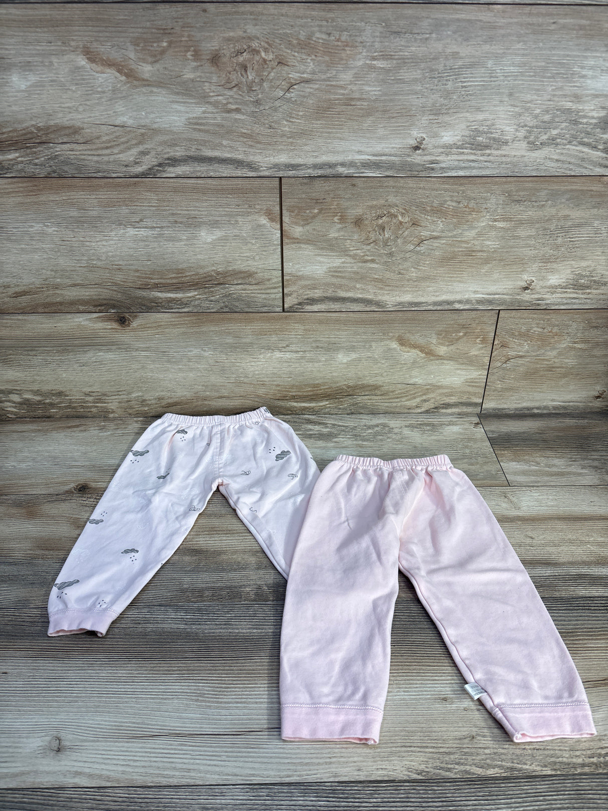 Flat lay of two baby pull-on pants, one solid pink and one pale pink with gray cloud print on wood background