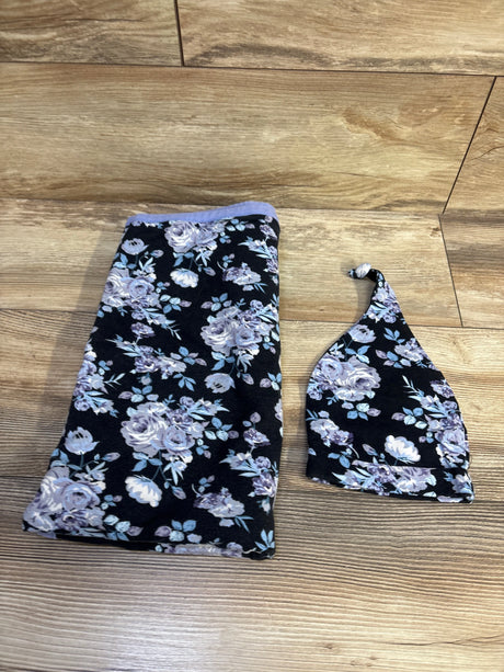 Flat lay of 2-piece floral swaddle blanket and knotted hat showing lavender and white roses on black fabric on wood floor