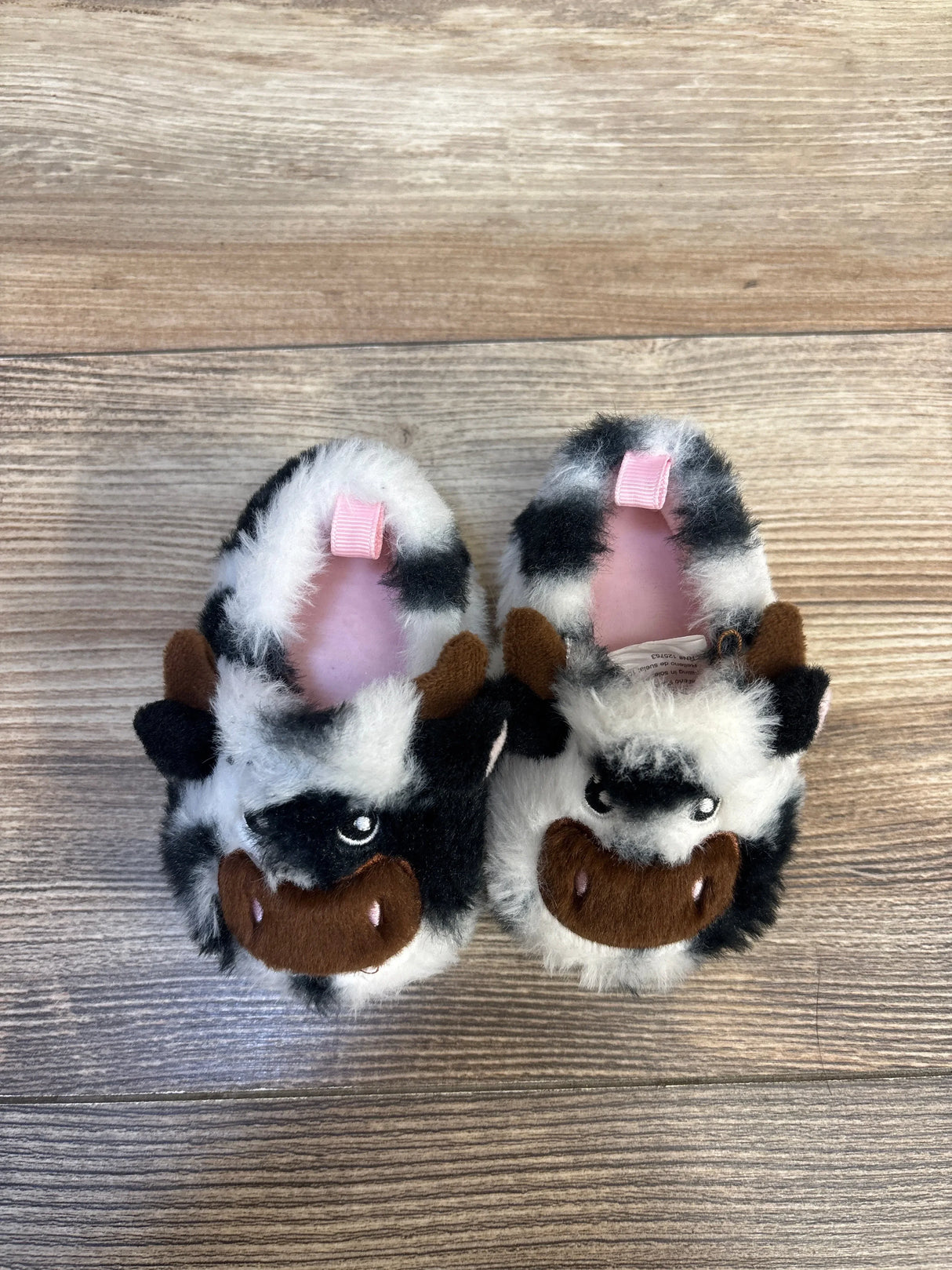 Plush baby slippers with cow faces, black and white fur, on wood background