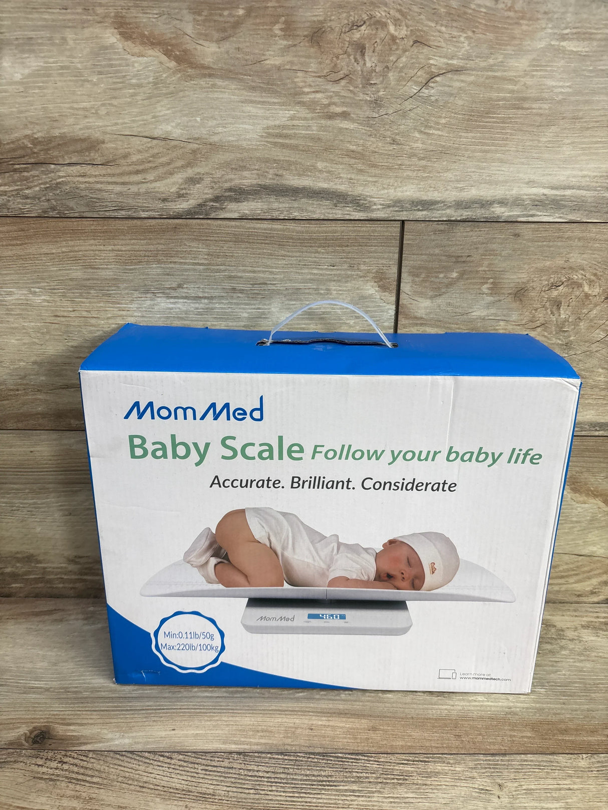 Front view of MomMed baby scale box showing sleeping baby on scale and weight range on wood background