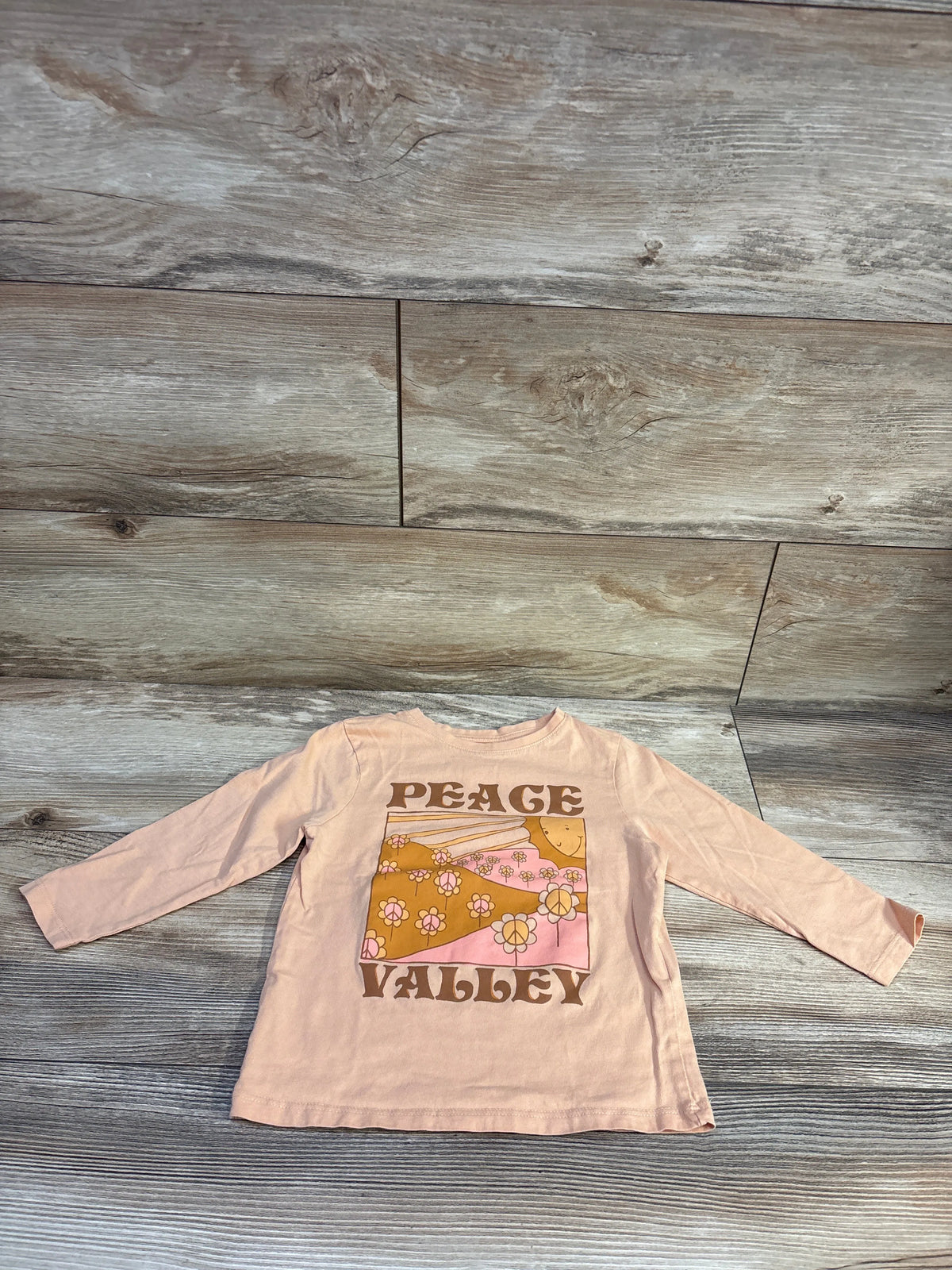 Front view of orange Cotton On Kids Peace Valley shirt showcasing long sleeves and floral Peace Valley graphic on a wood plank background