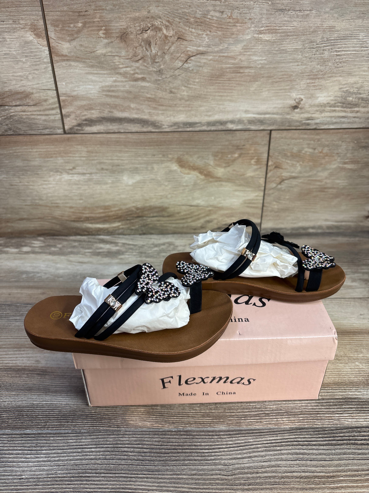 Three quarter view of black Flexmas toddler sandal showcasing rhinestone butterfly straps on a pink shoebox and wood floor