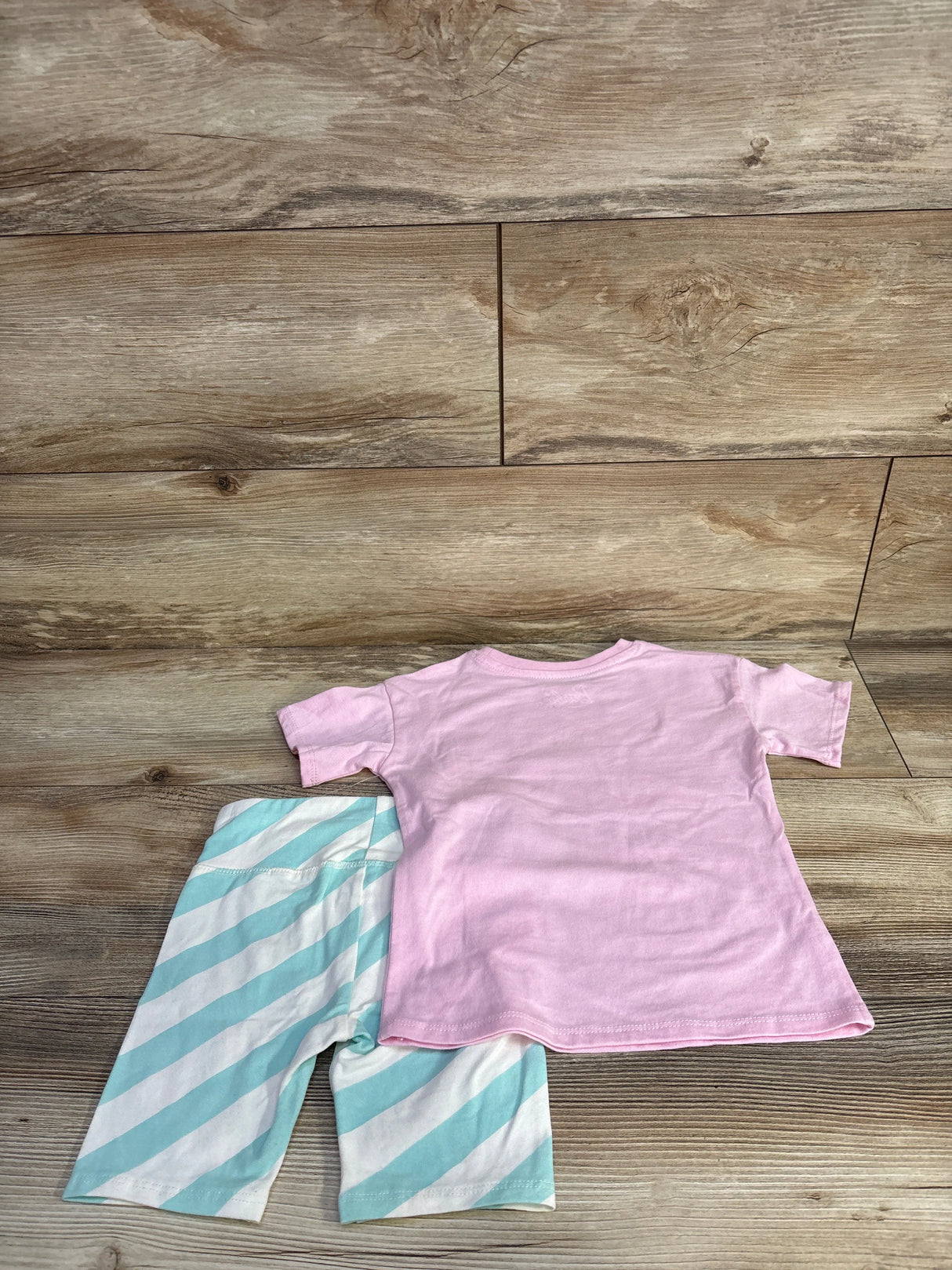 Flat-lay back view of Malibu Barbie 2-piece toddler set: pink T-shirt and mint-striped knee-length bottoms on wood background