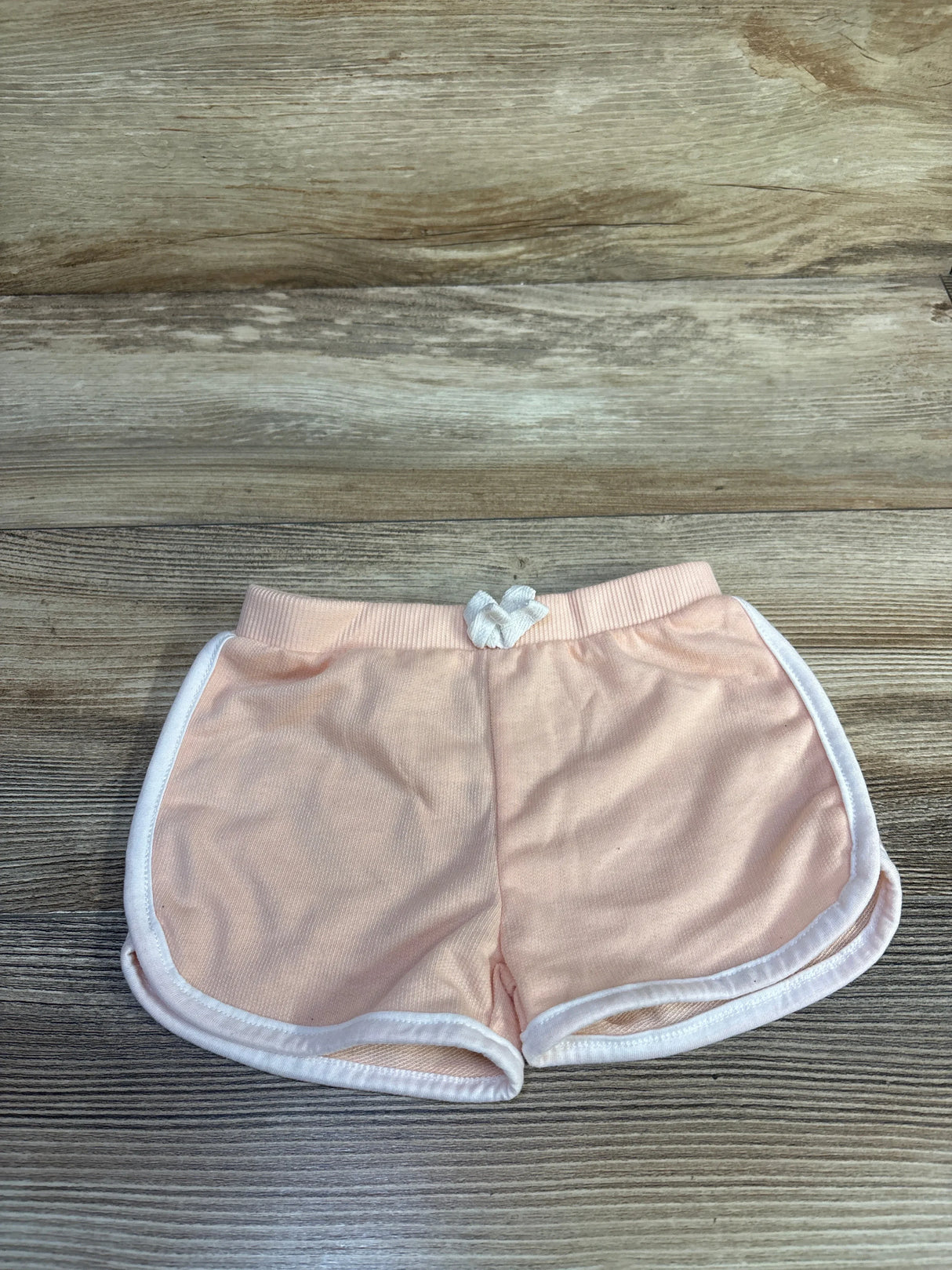Flat lay of pink toddler shorts with white piping and small front bow, elastic waistband visible, on wood background