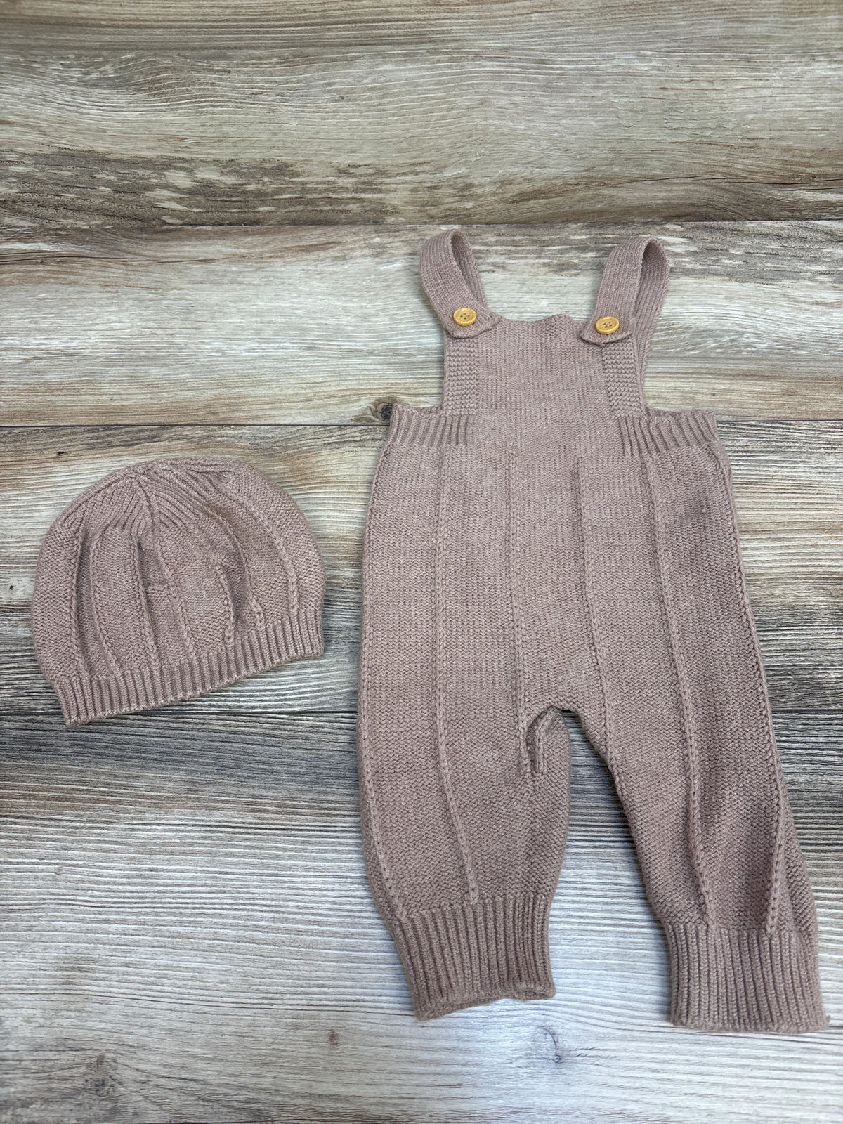 Top view of tan knit overalls and matching hat set showcasing buttoned straps and ribbed cuffs on a wood grain background