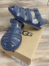 Front-and-side view of UGG Josiah Josiah Fisherman toddler sandals in navy showcasing woven leather straps, contrast white stitching, adjustable ankle strap, and rubber sole on a cardboard UGG box