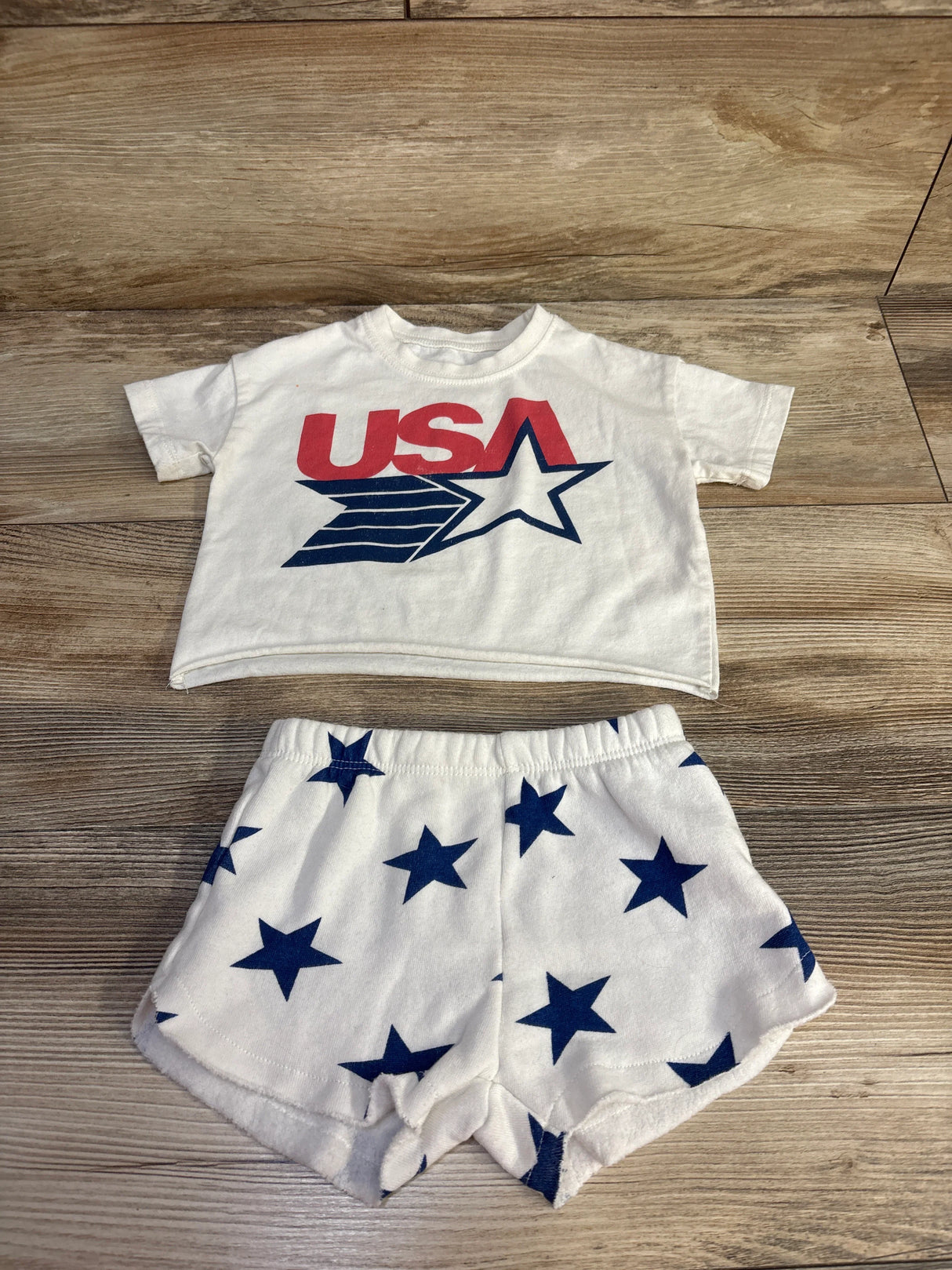 Flat lay of ivory 2-piece baby shirt and shorts set showing red USA graphic and navy star-print shorts on wood background