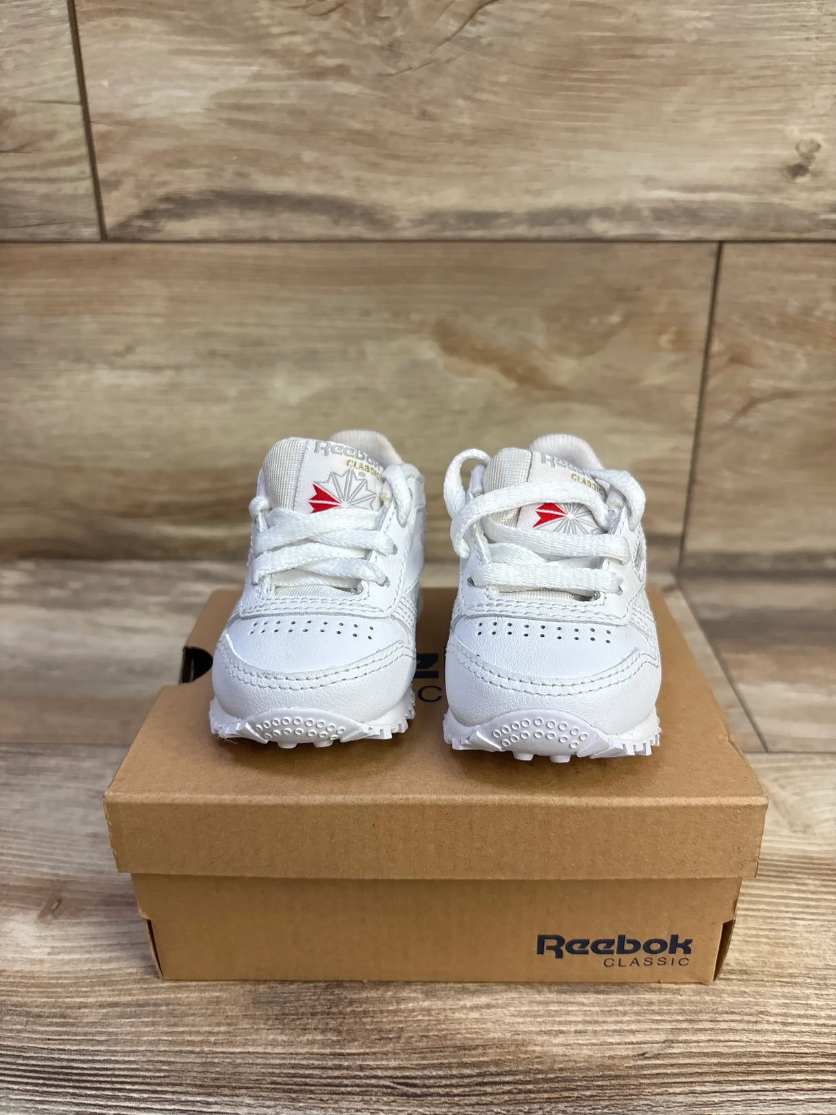 Front view of white Reebok Classic Leather infant sneakers (size 2C) showing lace-up leather upper, red logo on tongues, and textured rubber soles on a brown Reebok shoebox