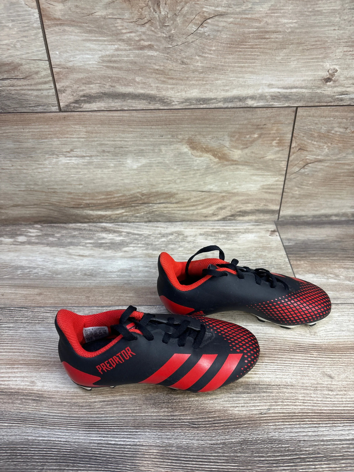 Top-down view of Adidas Predator 20.4 FxG toddler soccer cleats in black and red showing textured toe pattern, three red stripes and molded studs on wood floor