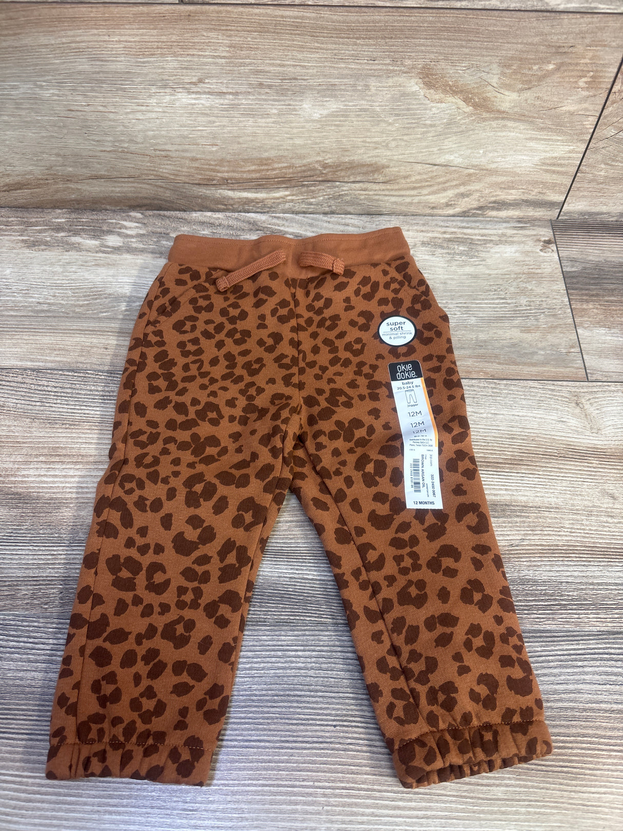 Flat lay of toddler brown leopard print jogger pants showing elastic waistband with drawstring, cuffed ankles, and 12M size tag on wood background