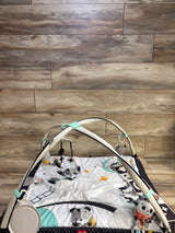 Top-front view of Tiny Love Magical Tales Black & White Gymini play gym showcasing padded black-and-white mat and crossed arches with hanging plush toys against wood-plank background