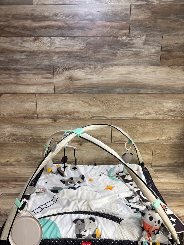 Top-front view of Tiny Love Magical Tales Black & White Gymini play gym showcasing padded black-and-white mat and crossed arches with hanging plush toys against wood-plank background