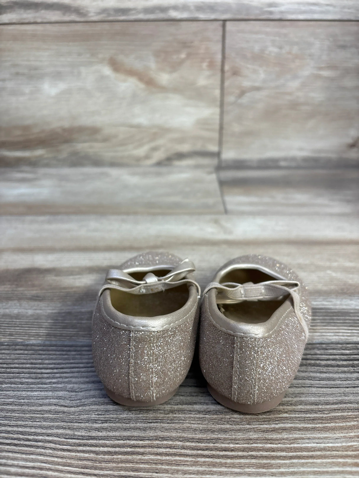 Rear view of Koala Kids gold glitter bow ballet flats showcasing glitter finish and elastic bow strap on wood-plank background