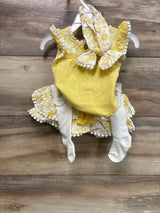Flat lay of yellow 4-piece outfit showing embroidered dress with pompom trim, floral headband and white tights on wood