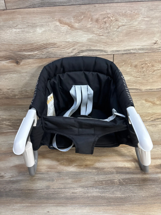 Black portable clip-on high chair with safety straps for babies, shown on wood floor