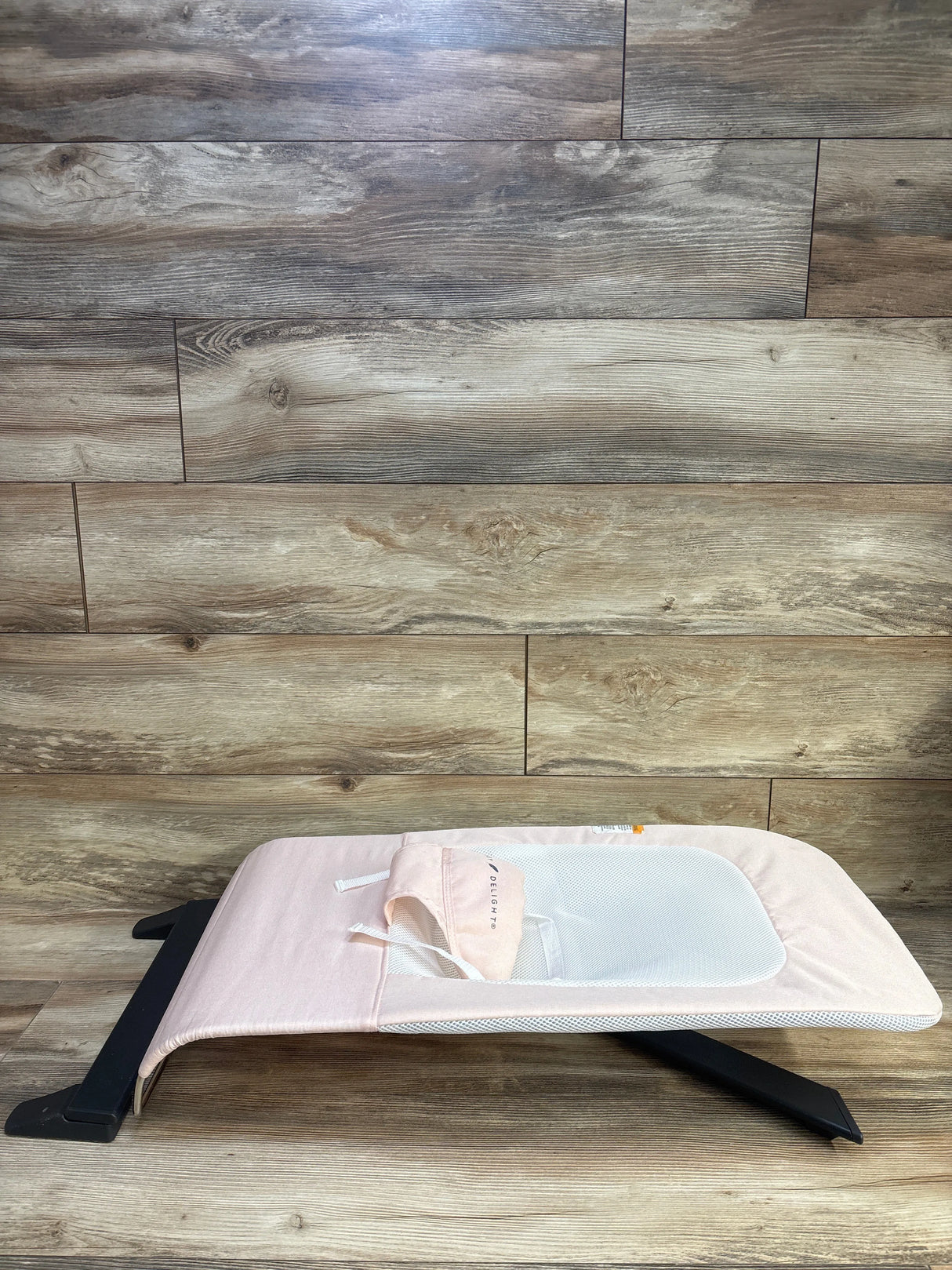 Side view of Baby Delight bouncer in light pink showing mesh seat and foldable black frame on wood floor
