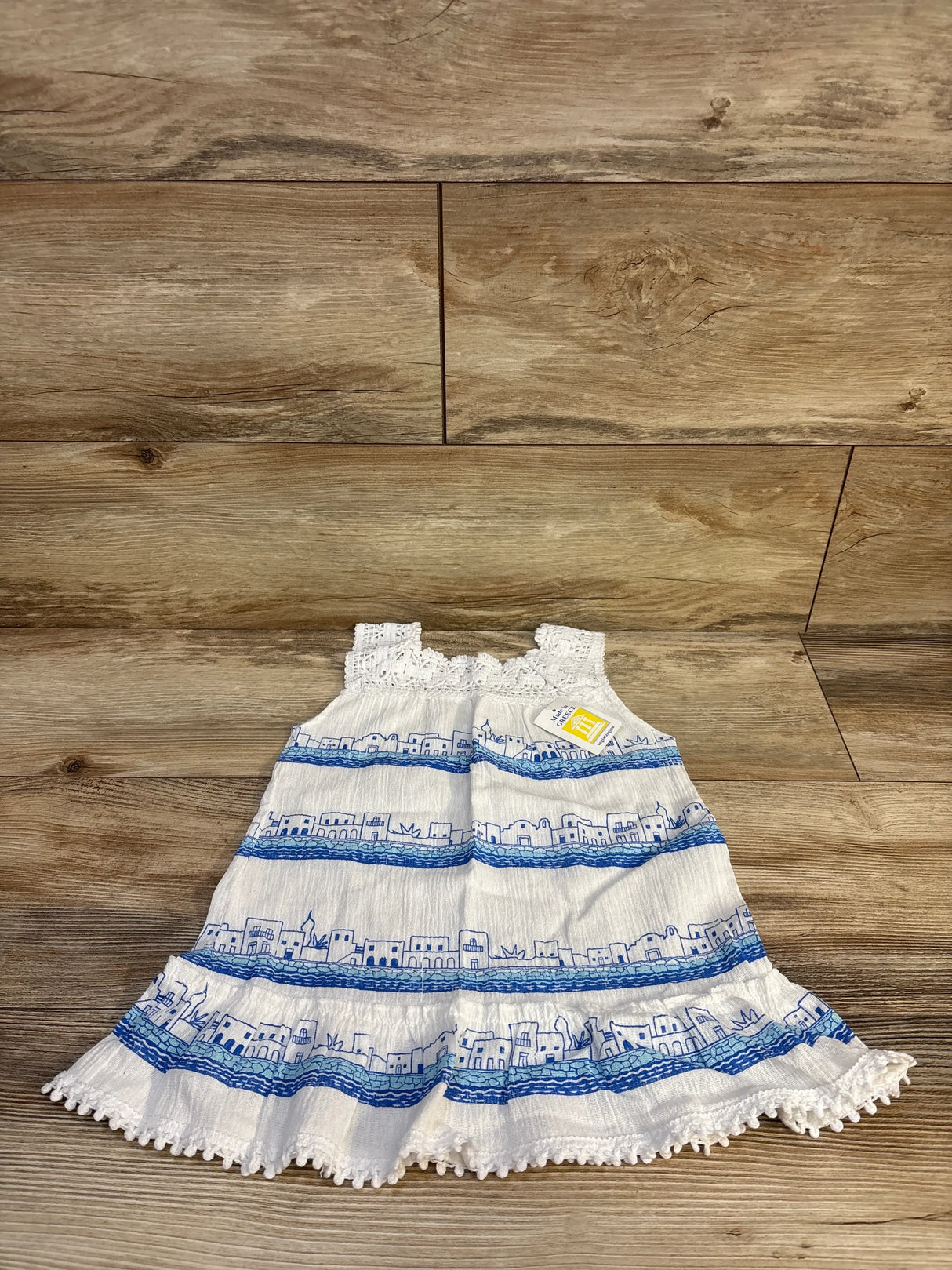 Front view of NEW Vogiatzoglou Kanataki Bebe dress white sz 2T showcasing sleeveless tiered dress with blue Greek village print and ruffle hem with hangtag on wood plank floor