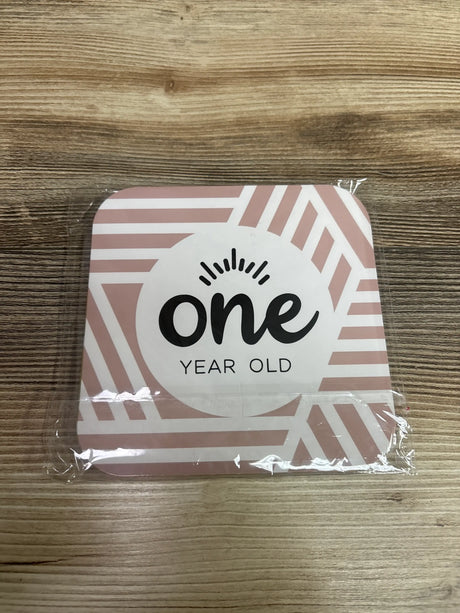 Front view of a 12pc monthly milestone photo prop card showing one year old text and pink geometric border on wood