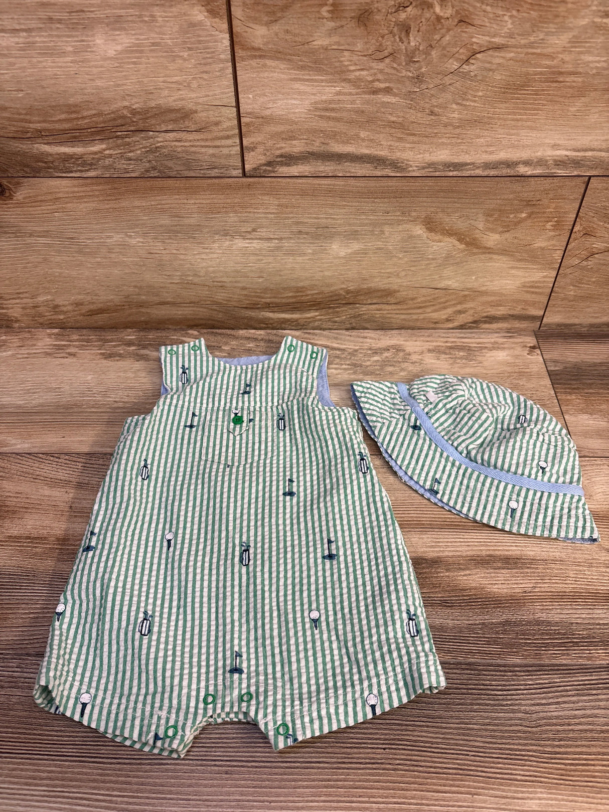 Front view of green seersucker baby romper and matching hat showing golf embroidered motifs and snap closures on wood floor