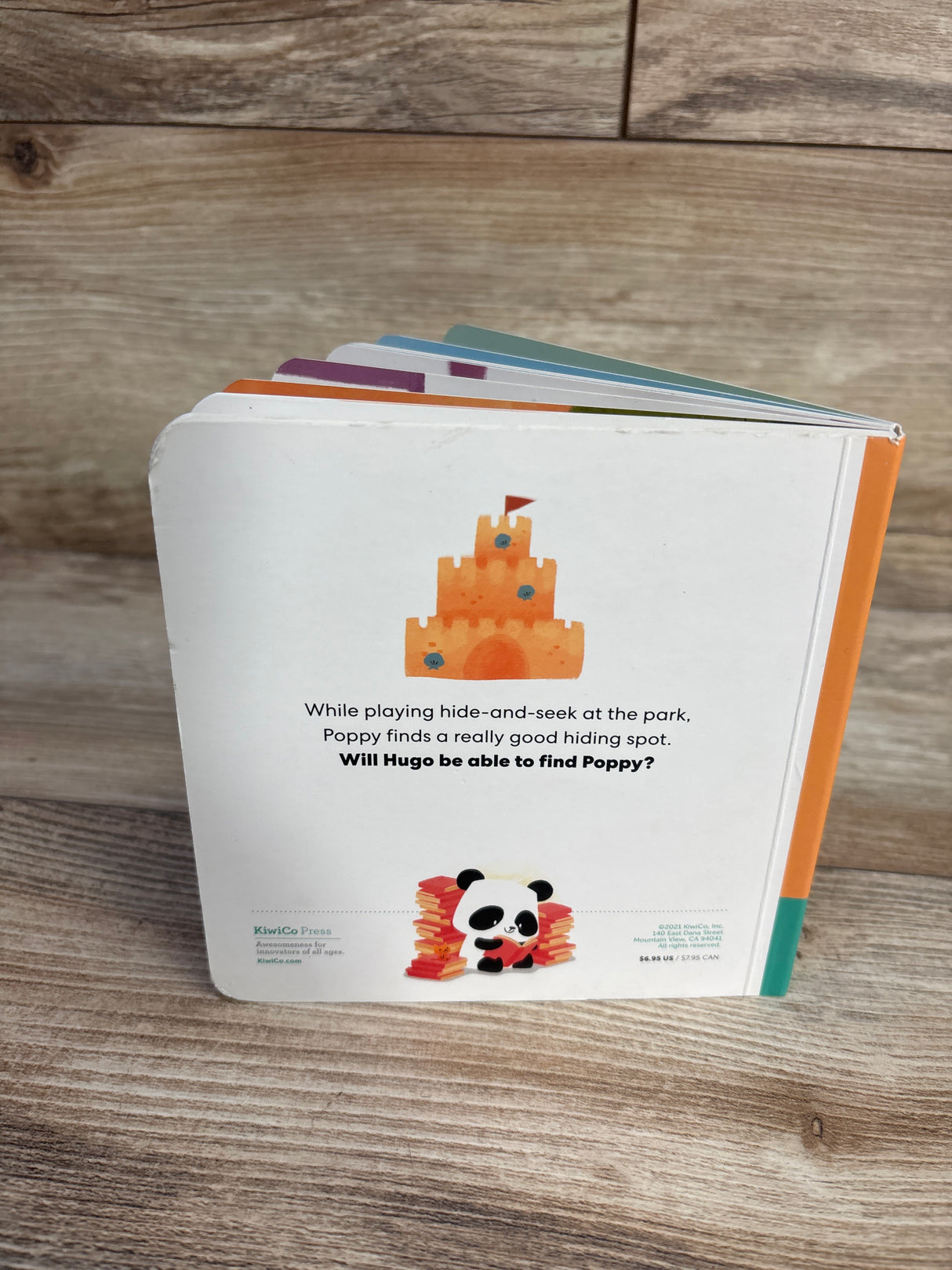 Angled back view of KiwiCo Where's Poppy board book showing fanned colorful board pages and rounded corners on wood surface