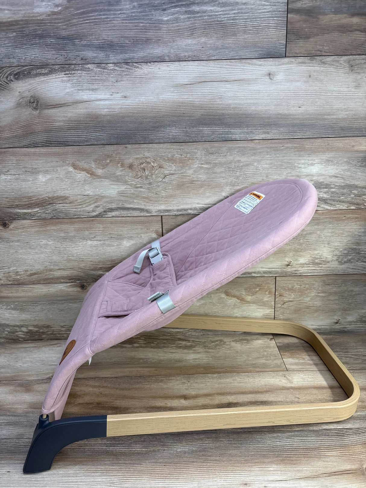 Side view of ANGELBLISS baby bouncer in pink with quilted seat, safety harness, and wooden curved base on wood background