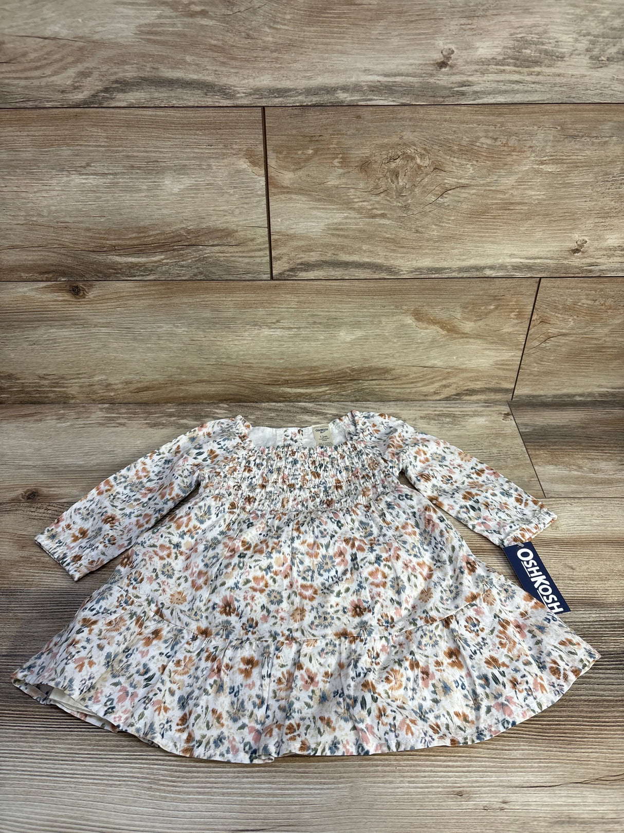 Flat lay of OshKosh smocked long-sleeve floral baby dress showing smocked bodice, ruffle hem and brand tag on wood background