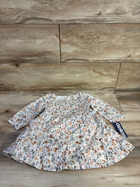Flat lay of OshKosh smocked long-sleeve floral baby dress showing smocked bodice, ruffle hem and brand tag on wood background