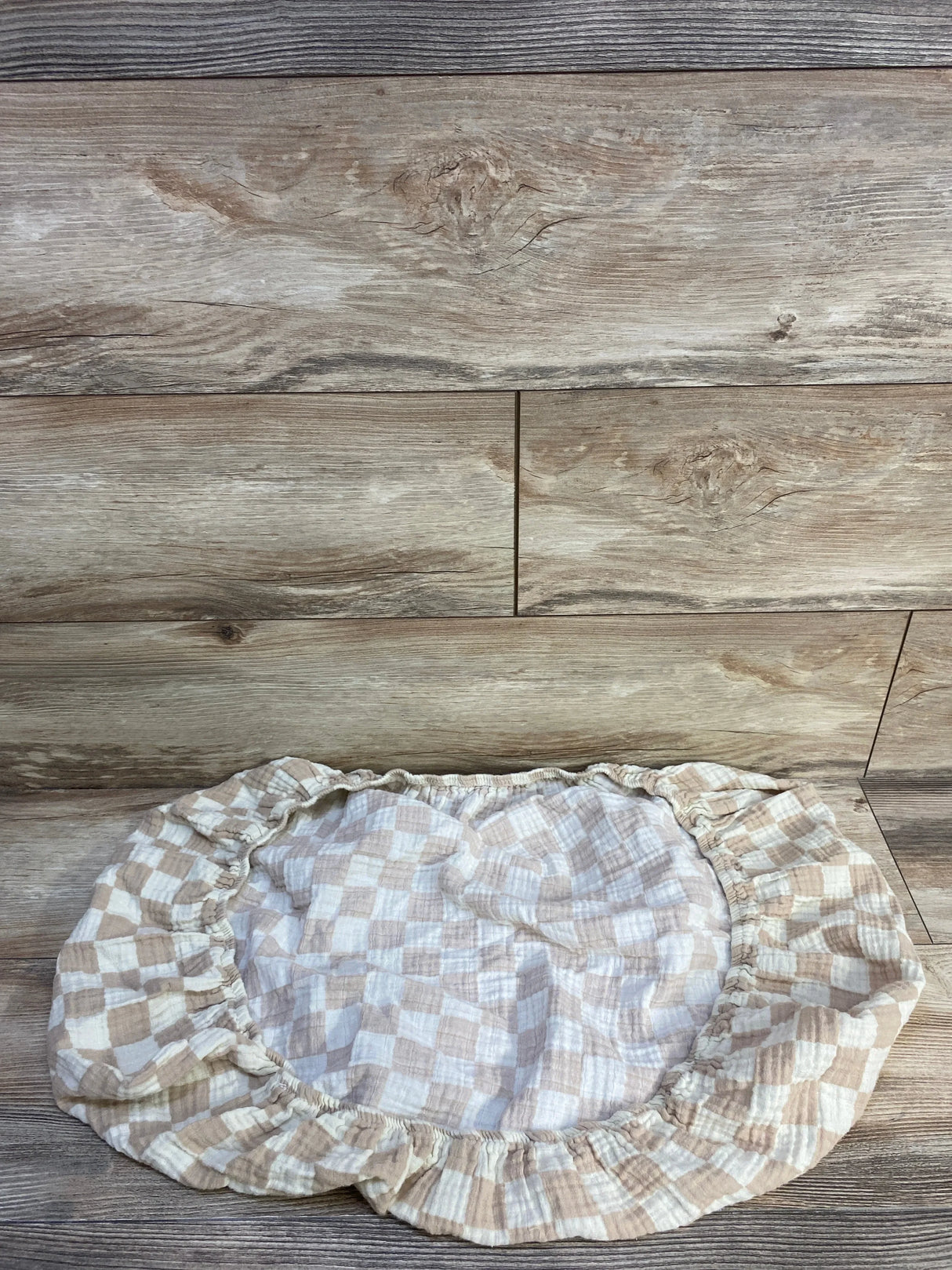 Top-down view of Brixton Phoenix muslin bassinet sheet with beige-and-white check and elastic fitted edge on wood background
