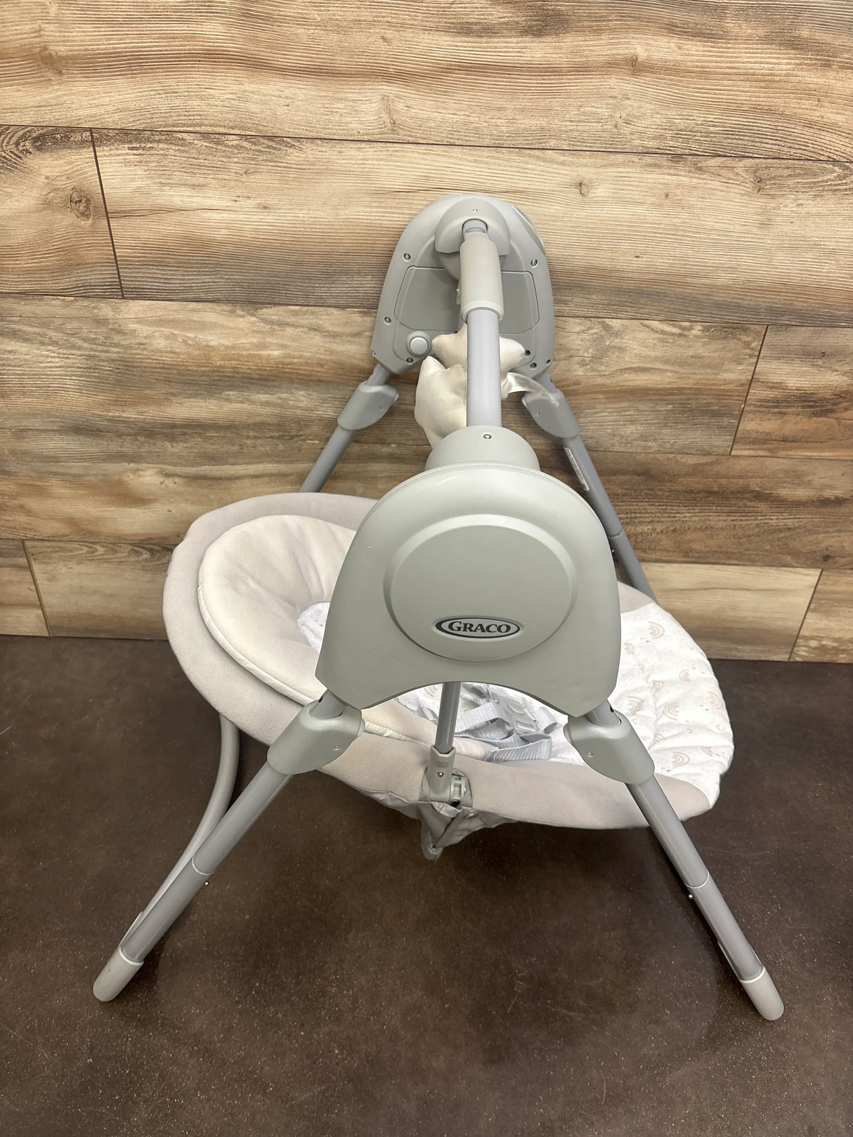 Rear-top view of Graco Slim Spaces baby swing with gray frame, plush beige seat and toy bar on wood background