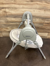 Rear-top view of Graco Slim Spaces baby swing with gray frame, plush beige seat and toy bar on wood background