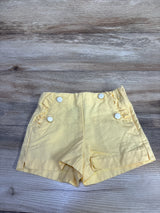 Top down view of Janie and Jack yellow shorts with four white buttons and ruffled pockets on wood background