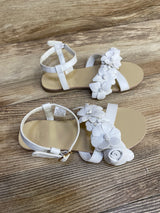 Top-down view of Janie and Jack white sandals with flower applique strap, ankle buckle, size 12 tag on wood floor