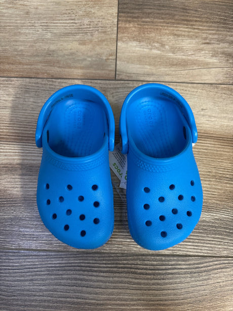 Top-down view of toddler blue Crocs Classic clogs showcasing toe ventilation holes and heel straps with wood floor background