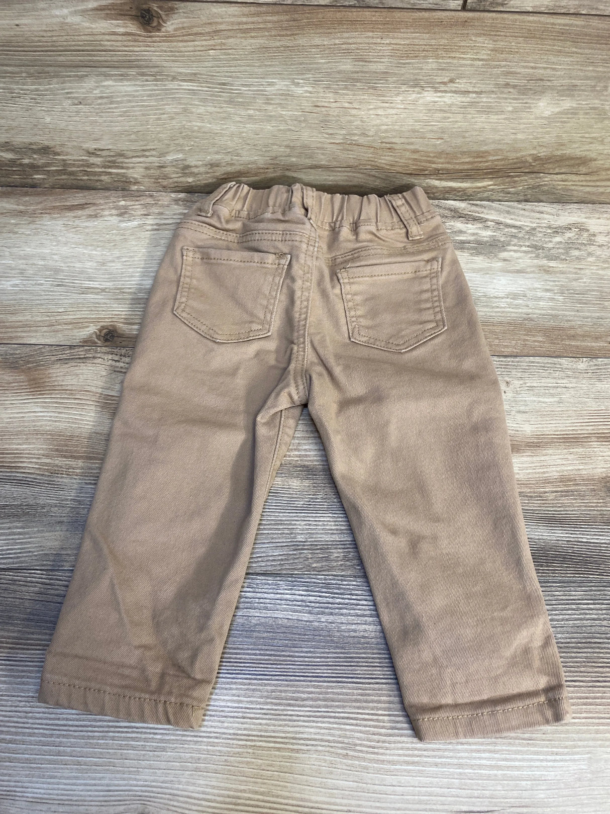 Back view of tan toddler pants with elastic waistband and two back pockets on wood surface