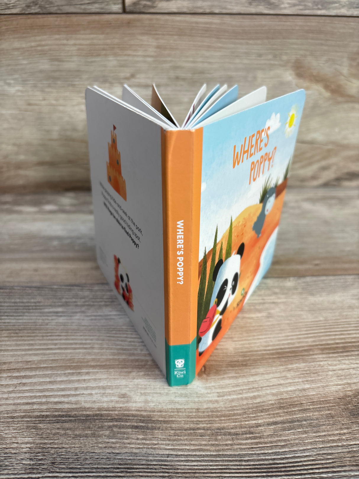 Angled front-right view of KiwiCo Where's Poppy board book showing orange spine, illustrated panda on cover, and fanned pages on wood surface