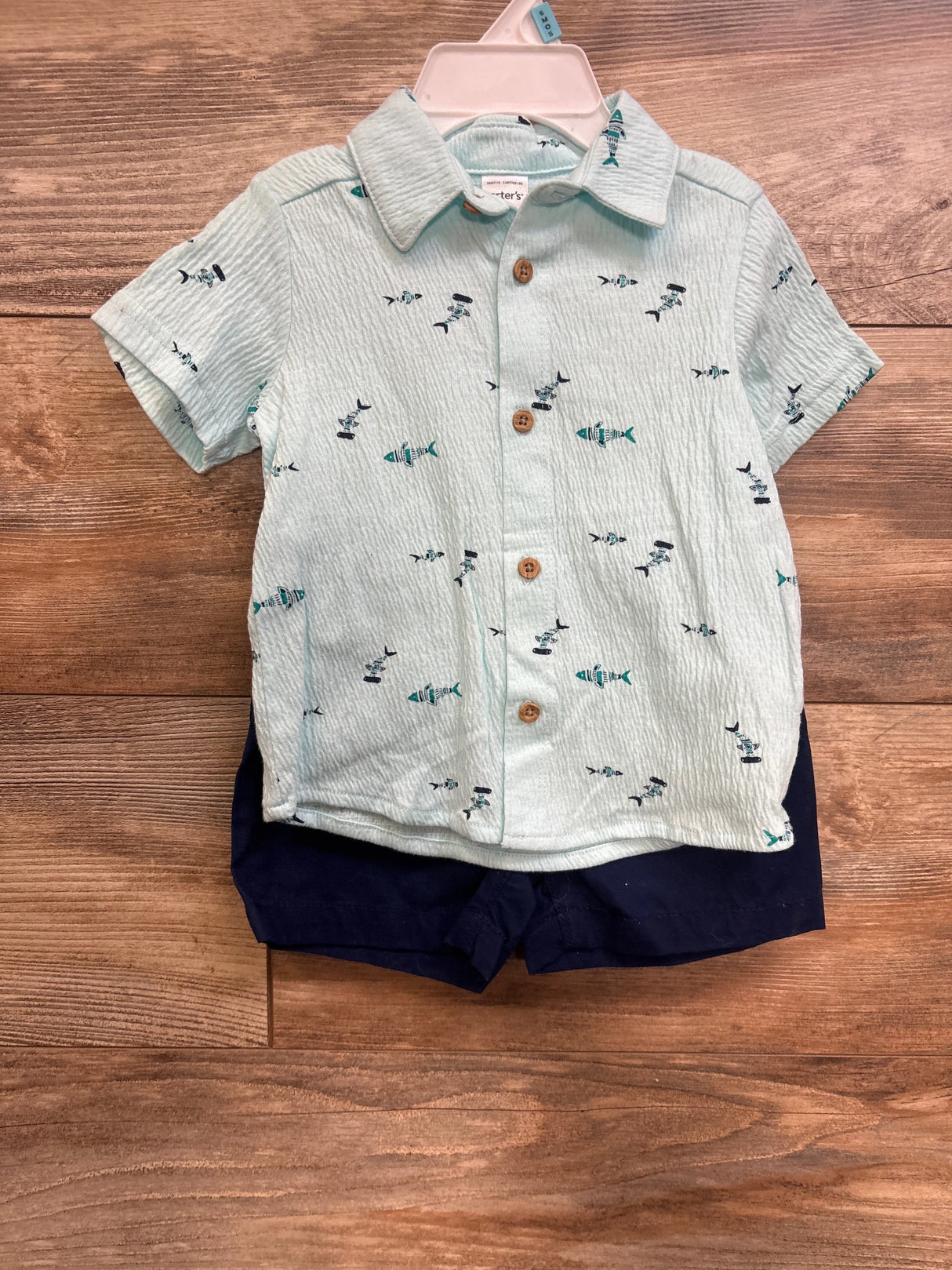 Front view of Carter's light-blue button-up shirt with fish print and wooden buttons with navy shorts on wood floor