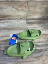 Top-down view of green Crayon Friends shark toddler slides size 9/10C showcasing shark-face strap, white teeth detail, and slip-on design on wood plank background