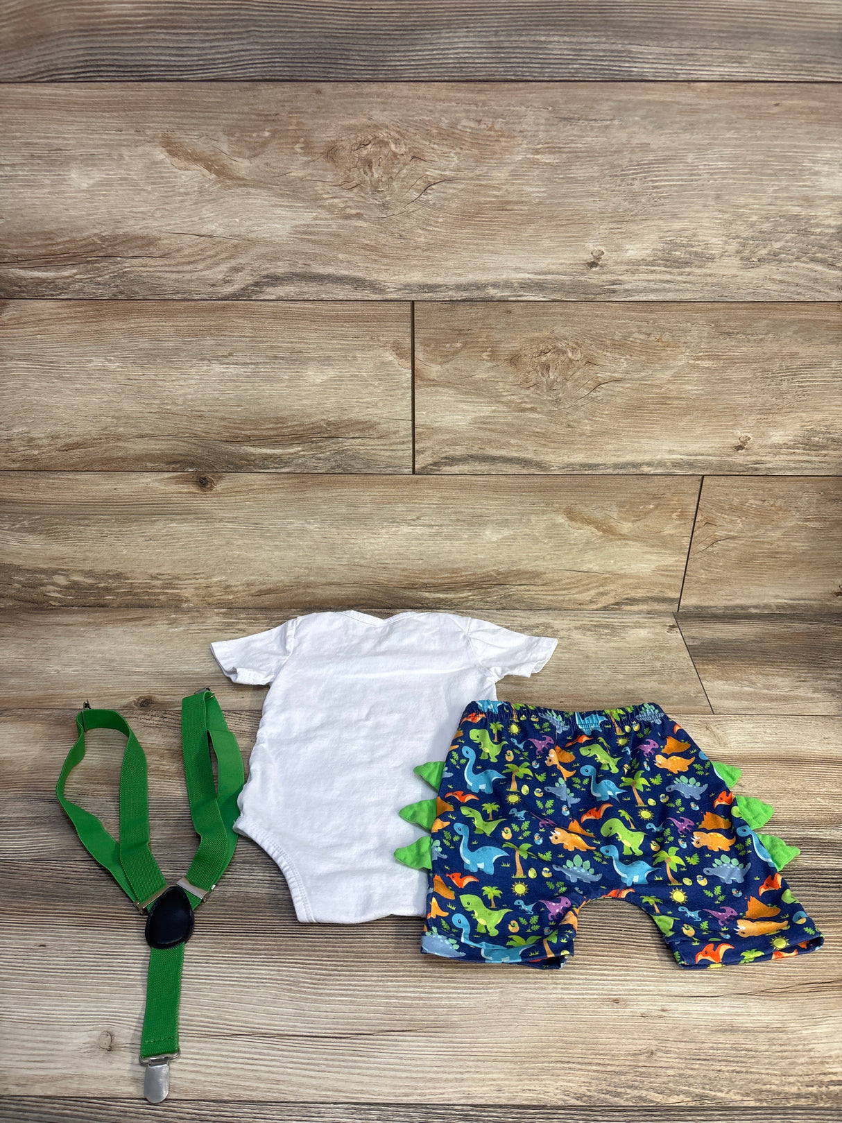 Flat lay of 3-piece baby dinosaur set: white onesie, navy dinosaur shorts with green spikes, green suspenders on wood