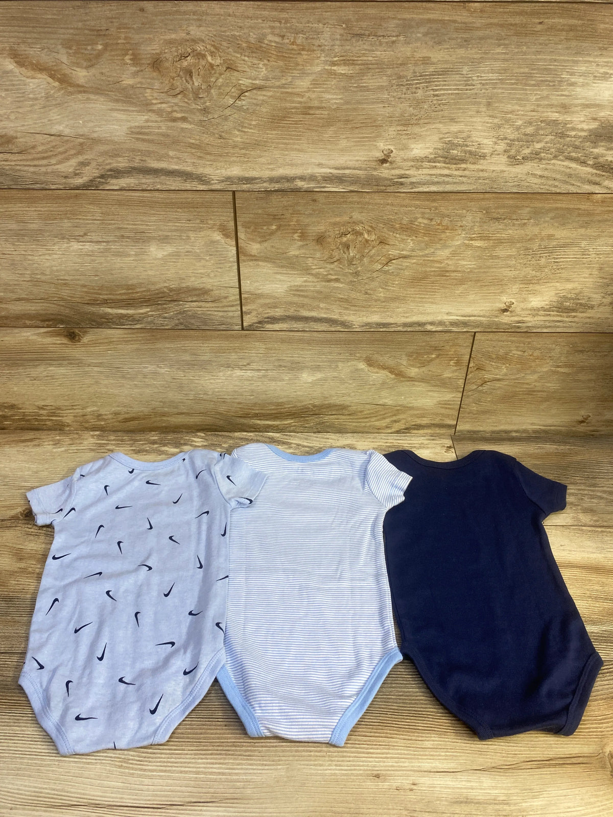 Front view of Nike three-pack baby bodysuits in light blue swoosh print, blue and white stripes, and navy on wood