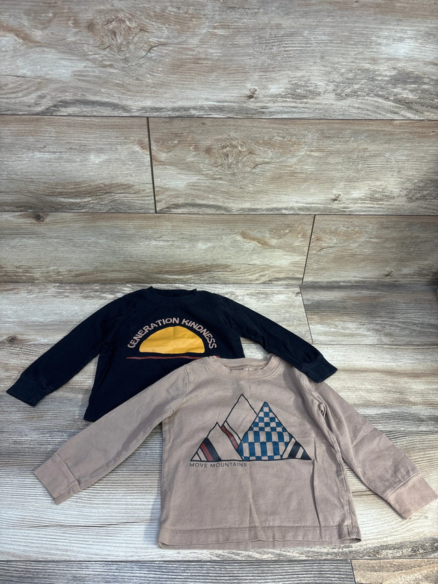 Flat lay of two toddler shirts (black, brown) showing Generation Kindness sun and Move Mountains designs on wood floor