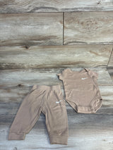 Flat lay of tan Nike 2-piece baby bodysuit and pants, size 6M, showing white Swoosh logos, elastic waistband and snap closures on light wood background