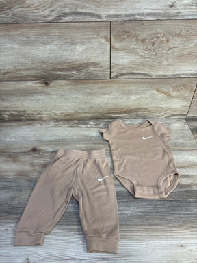 Flat lay of tan Nike 2-piece baby bodysuit and pants, size 6M, showing white Swoosh logos, elastic waistband and snap closures on light wood background