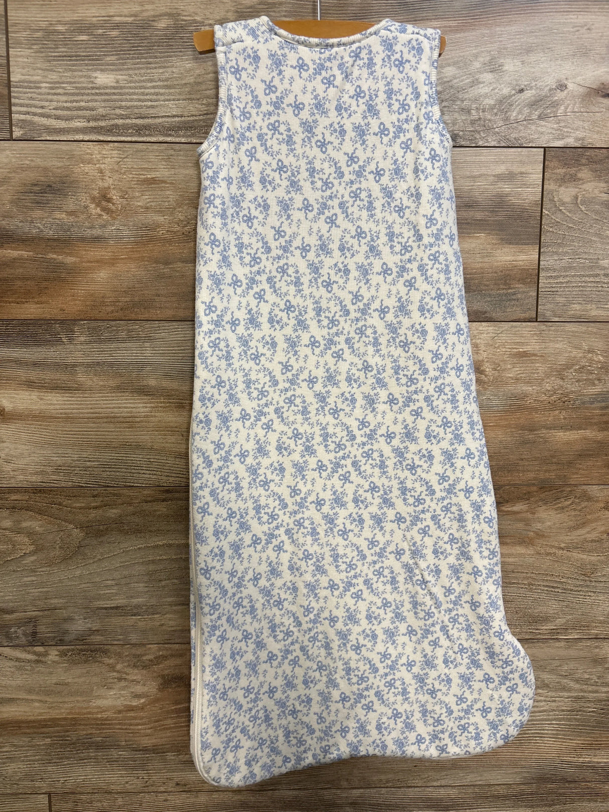 Flat lay of Angel Dear 1.0 TOG sleep bag showing sleeveless white knit with small blue floral-and-bow pattern on wood floor