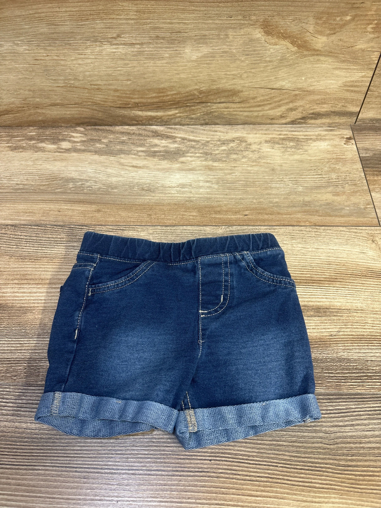 Flat lay of Jumping Beans blue pull on denim shorts size 18 months showing elastic waistband, rolled cuffs and front pockets on wood floor