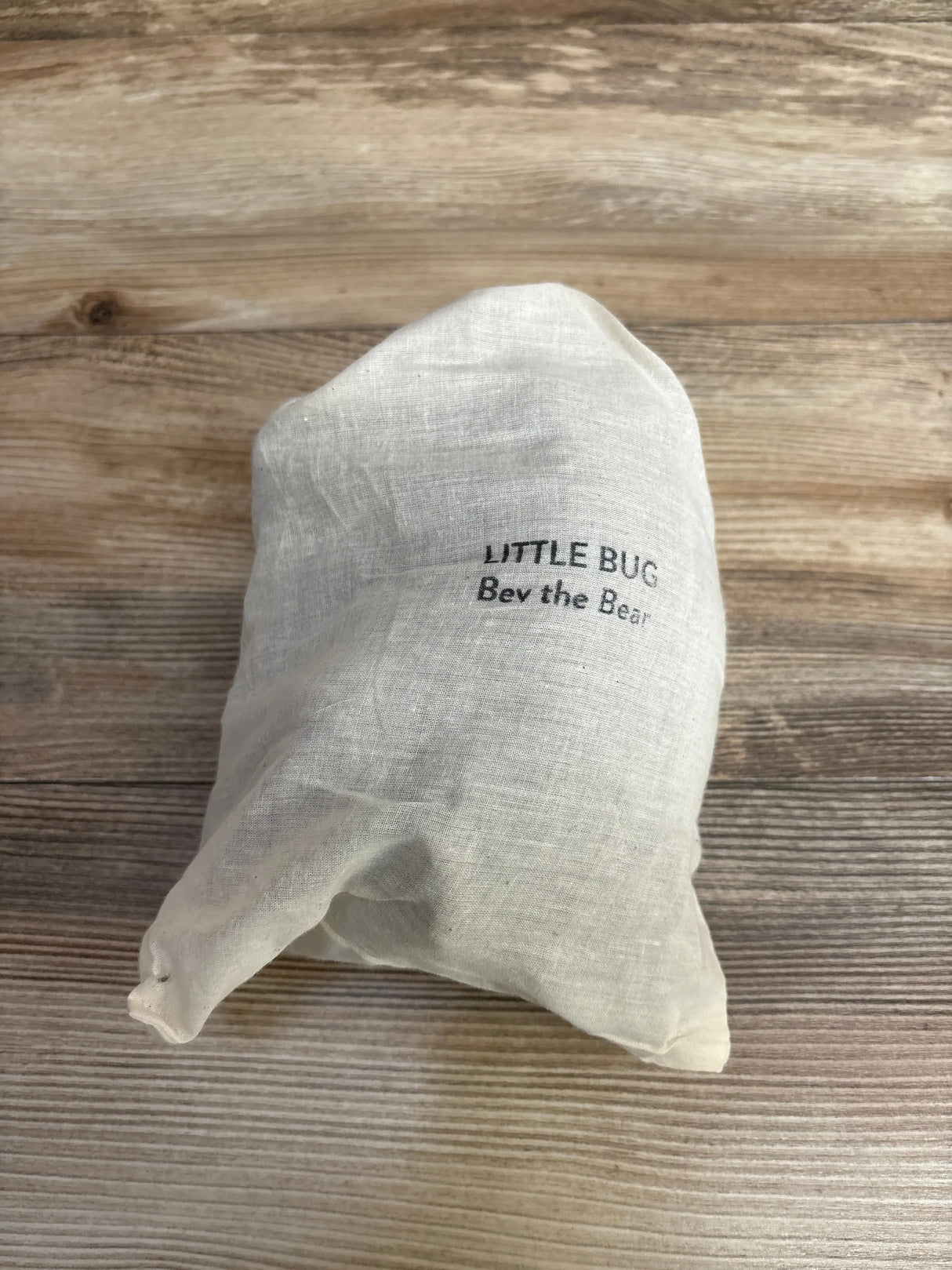 Flat lay of new Little Bug Bev the Bear lovie in cream linen drawstring pouch with printed brand label on wood plank floor