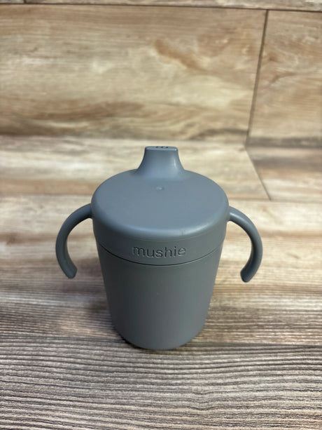 Front view of Mushie trainer sippy cup (7.7 oz) showcasing smoke grey silicone body, soft spout, two handles, and embossed mushie logo on wood tabletop background